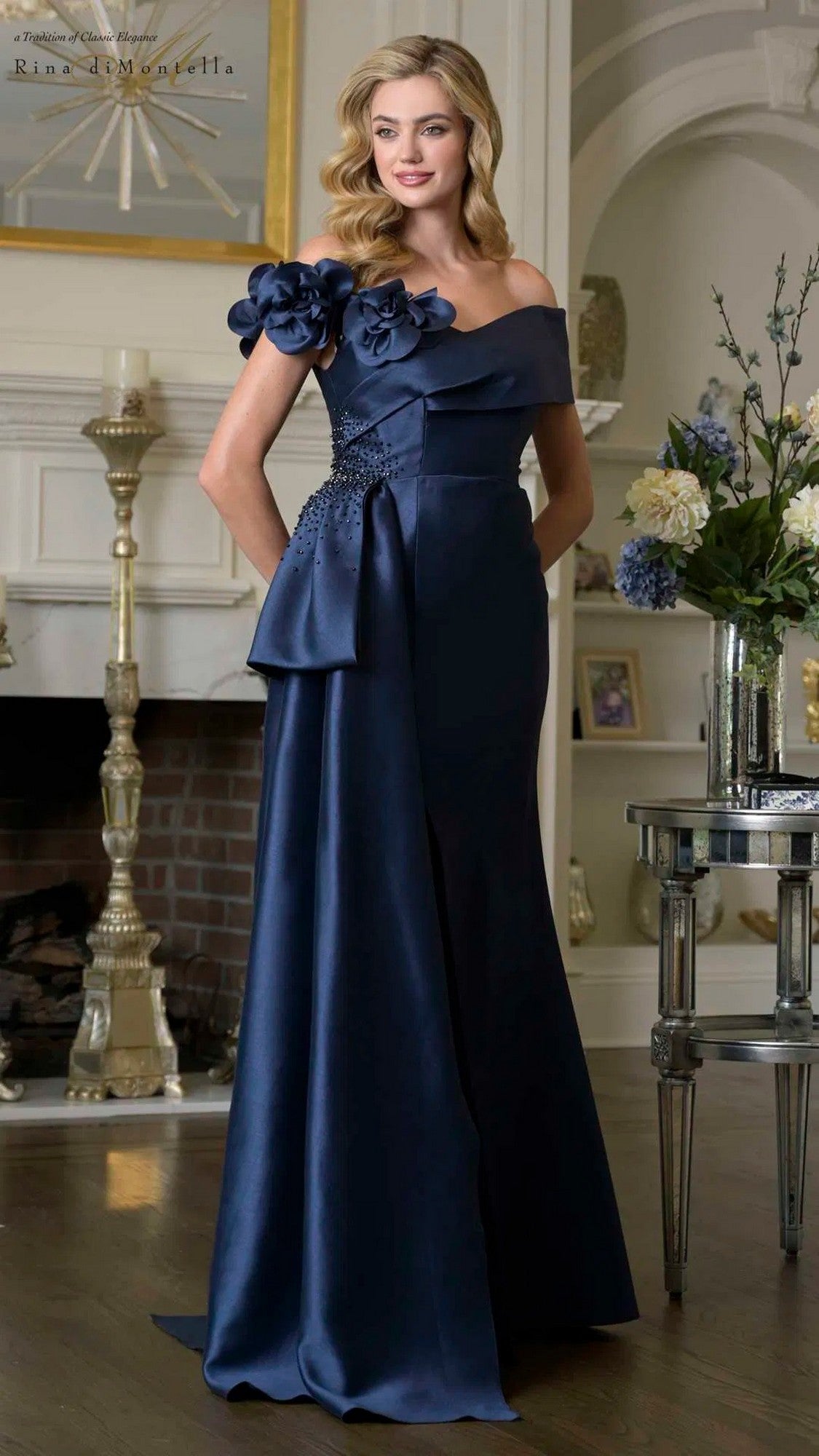 A woman stands in a stylish living room wearing the Rina Di Montella RD3407 long formal dress in navy, featuring an off-the-shoulder design with floral applique on one shoulder.